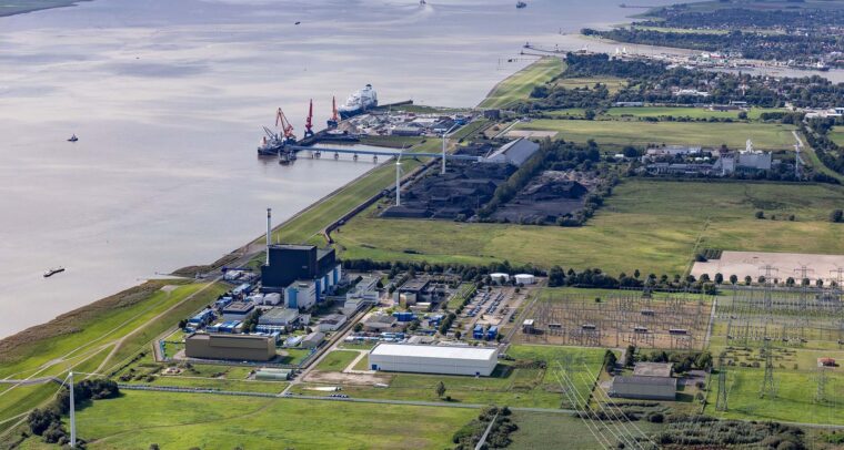 Vista diurna de la terminal flotante de GNL en Brunsbüttel (Alemania), donde el mar se encuentra con extensas áreas verdes. Se observa un puerto con buques y grúas, una planta industrial, turbinas eólicas y una subestación eléctrica, parte de la infraestructura energética del país. Rusia