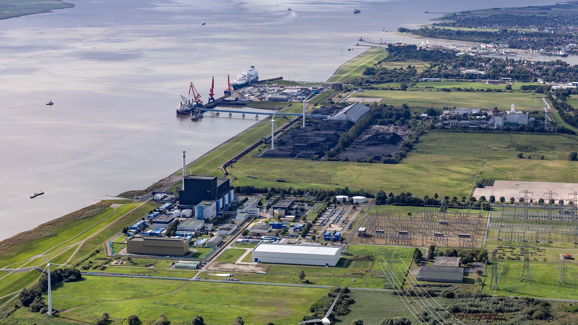 Daytime view of the floating LNG terminal in Brunsbüttel (Germany), a port featuring ships, cranes, an industrial plant, wind turbines, and an electrical substation, where the sea meets vast green areas. Russia