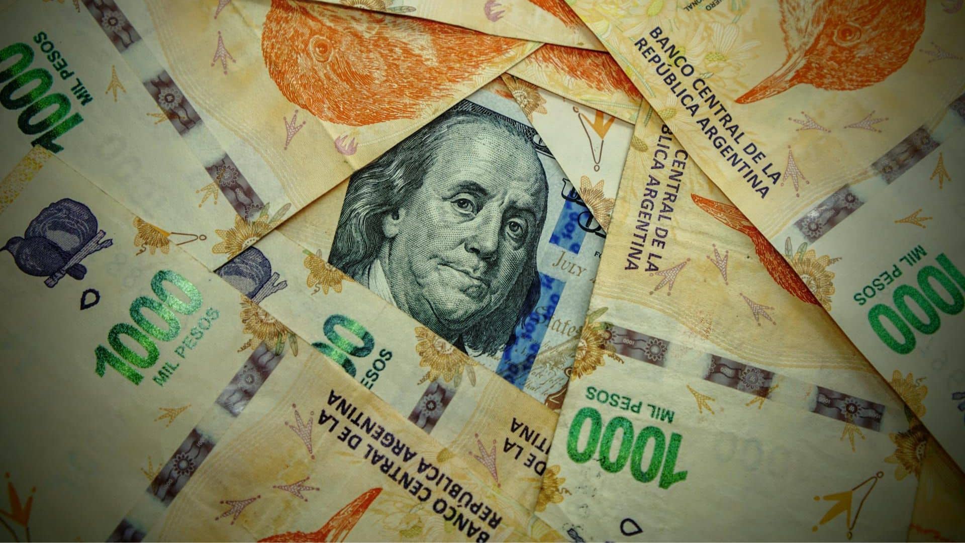 Detail of a green and black 100 US dollar banknote partially visible beneath several orange, violet, and green 1,000 Argentine peso notes. Argentina