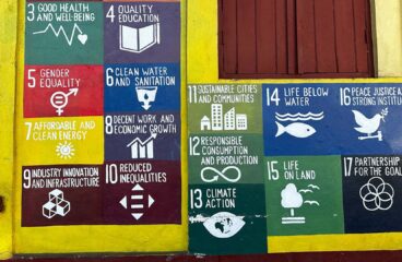 Mural pintado en una pared que representa los 17 Objetivos de Desarrollo Sostenible (ODS) de las Naciones Unidas, cada uno con un ícono y color distintivo. A la izquierda del mural hay una puerta roja con un cartel que dice "NO VENDING". A la derecha, sobre el mural, se encuentra una ventana de madera color terracota, enmarcada por una franja amarilla. Desarrollo